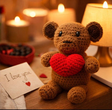 Crocheted monkey with a red heart and 'I love you' card on a wooden surface with candles in the background.