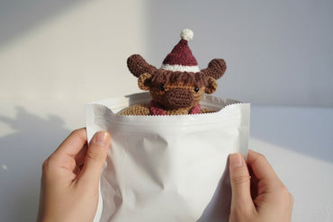 Handmade crochet cow toy peeking out from a white paper bag against a plain background