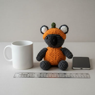 Crocheted toy with an orange outfit and hat on a white surface with a mug, phone, and ruler.
