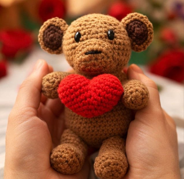 Hand holding a small brown monkey with a red heart, against a blurred floral background.