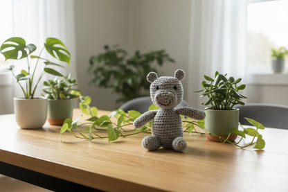 Crocheted hippo on a wooden table with potted plants in a bright room.