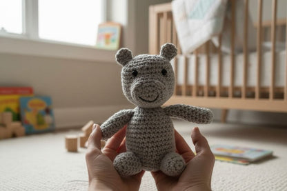 Hand holding a small crocheted hippo in a nursery setting with a crib and books in the background.