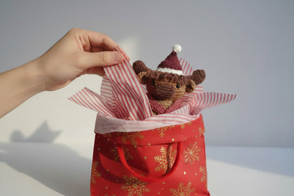 Hand holding a small brown cow in a red gift bag with snowflake pattern on a light background