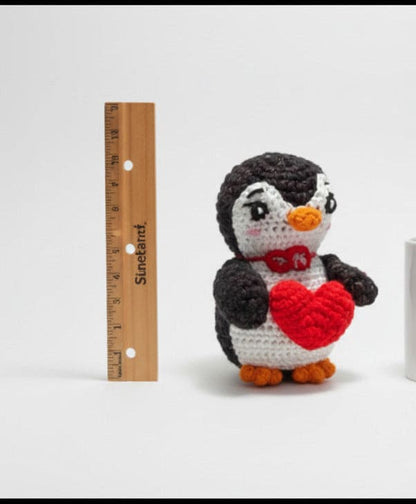 Crocheted penguin with a heart, a mug, and a ruler on a white background