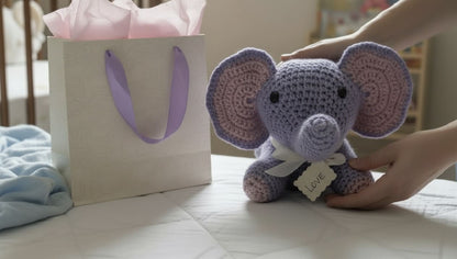 Handmade crochet elephant toy held by a person next to a gift bag with a purple ribbon.