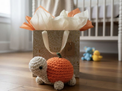 Crocheted turtle with pumpkin shell in front of a gift bag on a wooden floor.