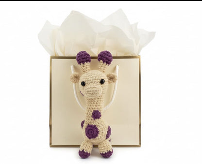  "Crocheted giraffe with purple spots in front of a gift bag."